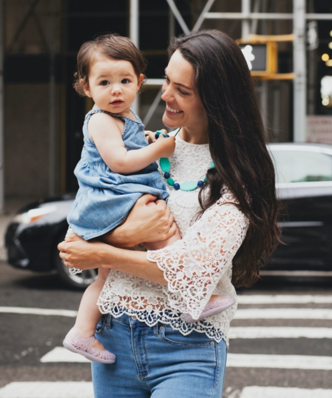 A woman with light skin tone and long dark hair is holding a baby in a cityscape. The baby is pulling on the turquoise Tribeca Necklace.