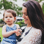 A woman with light skin tone and long dark hair is holding a baby while wearing the black Tribeca Necklace.