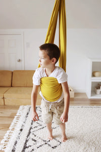 A child with light skin tone and short brown hair has the Retro Pineapple Sensory Swing stretched around their torso with their feet on the ground.