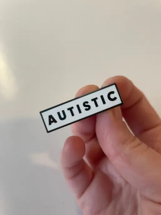 A hand holds up the Autistic Enamel Pin.