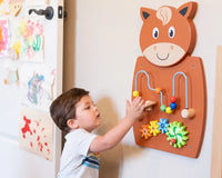 A child with light skin tone and short brown hair moves the wooden beads on the Horse Activity Wall Panel.