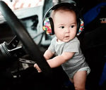 A baby wears the BANZ Baby Noise Reducing Earmuffs while seated behind a steering wheel.