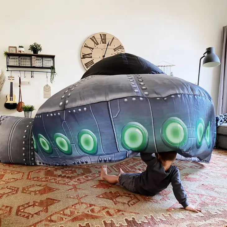 The UFO AirFort is set up in a living room. A child with light skin tone is sitting on the floor with their legs spread out in front of them, inside the tent. They are lifting up the side of the tent with one arm and leaning back on the other.