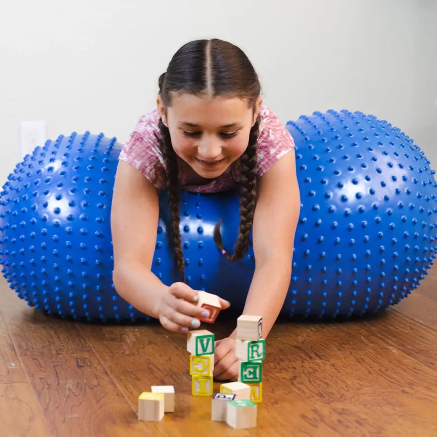 A child lays in the middle of the Peanut Stability Ball on her belly while playing with blocks.