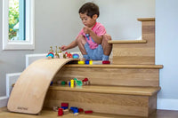 A child with medium-light skin tone and short brown hair is squatting on a stair landing. There is a Kinderboard Balance Board upsidedown sitting next to them, and several toys are sitting at the top of the board.