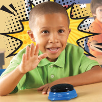 A child with medium-dark skin tone and very short black hair is smiling while their hand is posed above the blue Recordable Answer Buzzer.