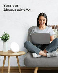 A person with light skin tone and long dark brown hair is sitting criss-cross applesauce with a laptop on their legs. The Light Therapy Lamp is illuminated on a small wooden table next to a vase with a green plant.
