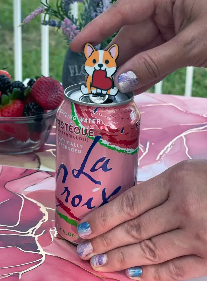 A hand pops a soda tab with the Corgi Soda Tab Buddy.