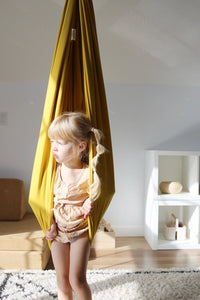 A child with light skin tone stands on a carpet with a Retro Pineapple Sensory Swing covering pulled around their torso.