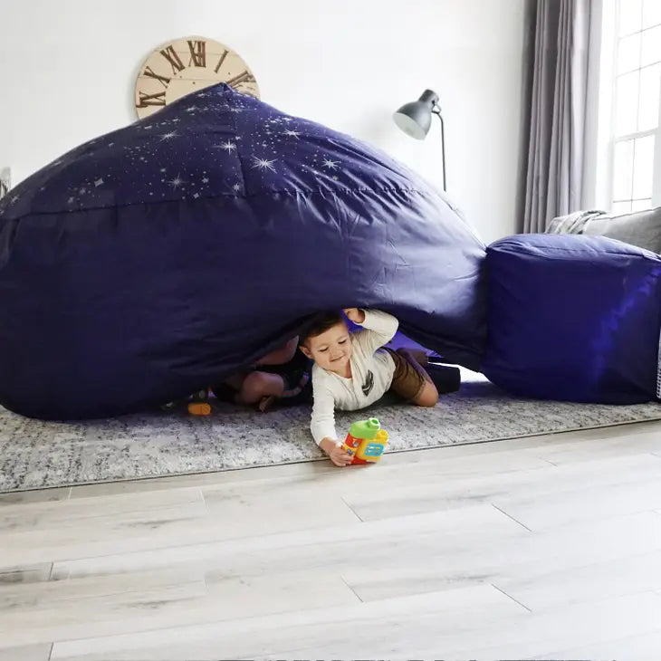 A child with light skin tone is on their belly, partially under the Starry Night AirFort. They are holding up the side of the tent and holding onto a toy with their other arm, which is extended out of the tent.