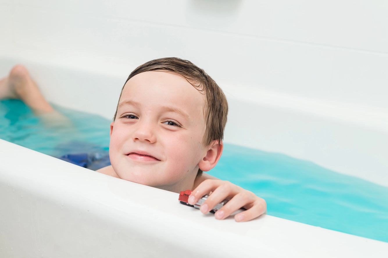 A child with light skin tone and brown, wet hair peaks their chin up over the edge of the tub while they balance a train on the ledge.