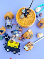 A mining cart sits at the bottom of the picture next to a plastic bat figurine. There is an open container of Prickly Pear scented dough with a crystal spider figurine on top.