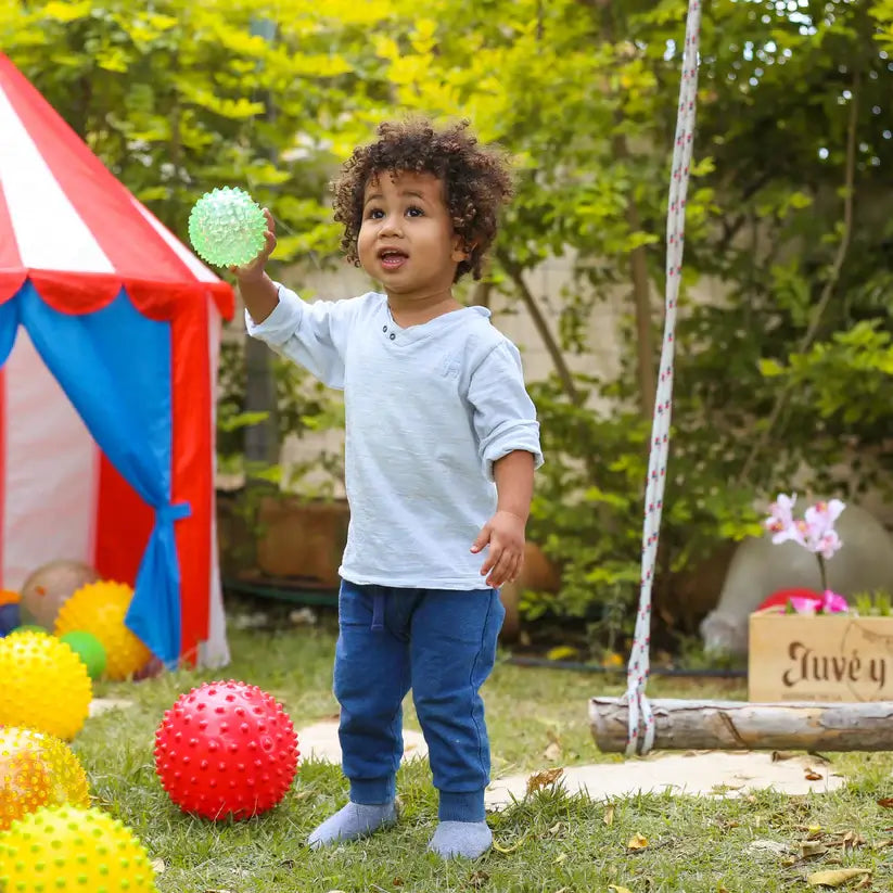 A child stands in a yard and holds up the green Original Sensory Balls See-Me.