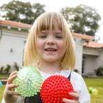 A child smiles into the camera and holds up two Original Sensory Balls.