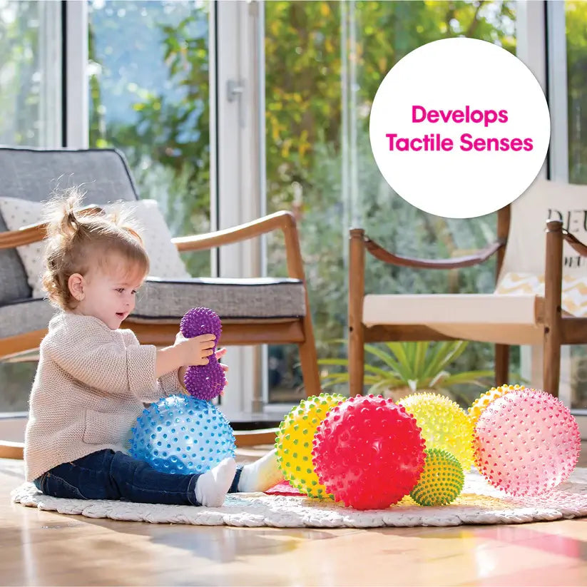 A baby holds a purple spiky peanut shaped ball on a carpet full of Sensory Shapes & Balls.