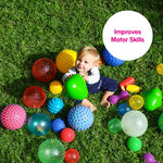 A baby on the grass holds up a green triangle ball while surrounded by a large number of sensory balls.