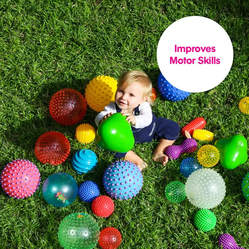 A baby on the grass holds up a green triangle ball while surrounded by a large number of sensory balls.
