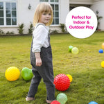 A child wearing black overalls stands in the grass and looks behind them at the camera while surrounded with Edushape sensory balls.