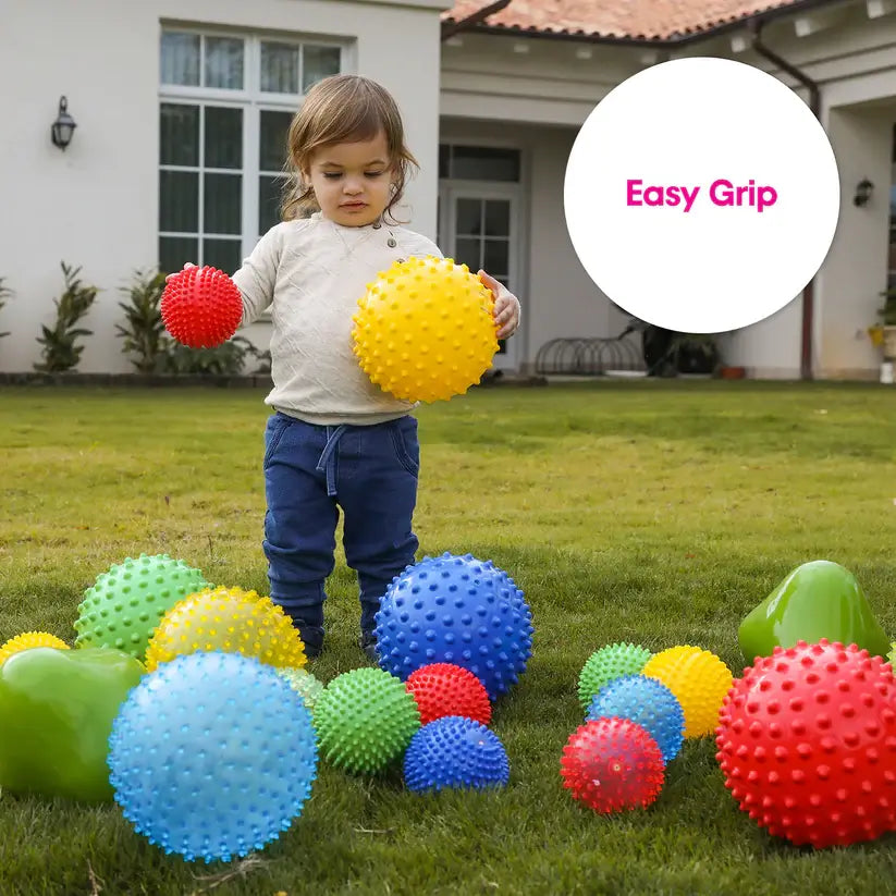 A toddler stands on grass and holds up a small red spiky ball and a large yellow spiky ball.