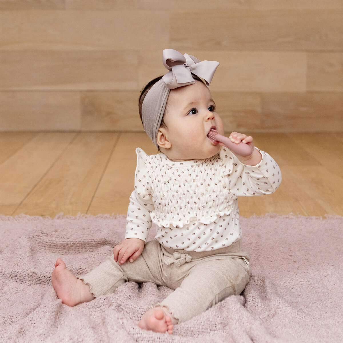 A toddler sits up and puts the Blush ezpz Baby-Led Toothbrush in its mouth.