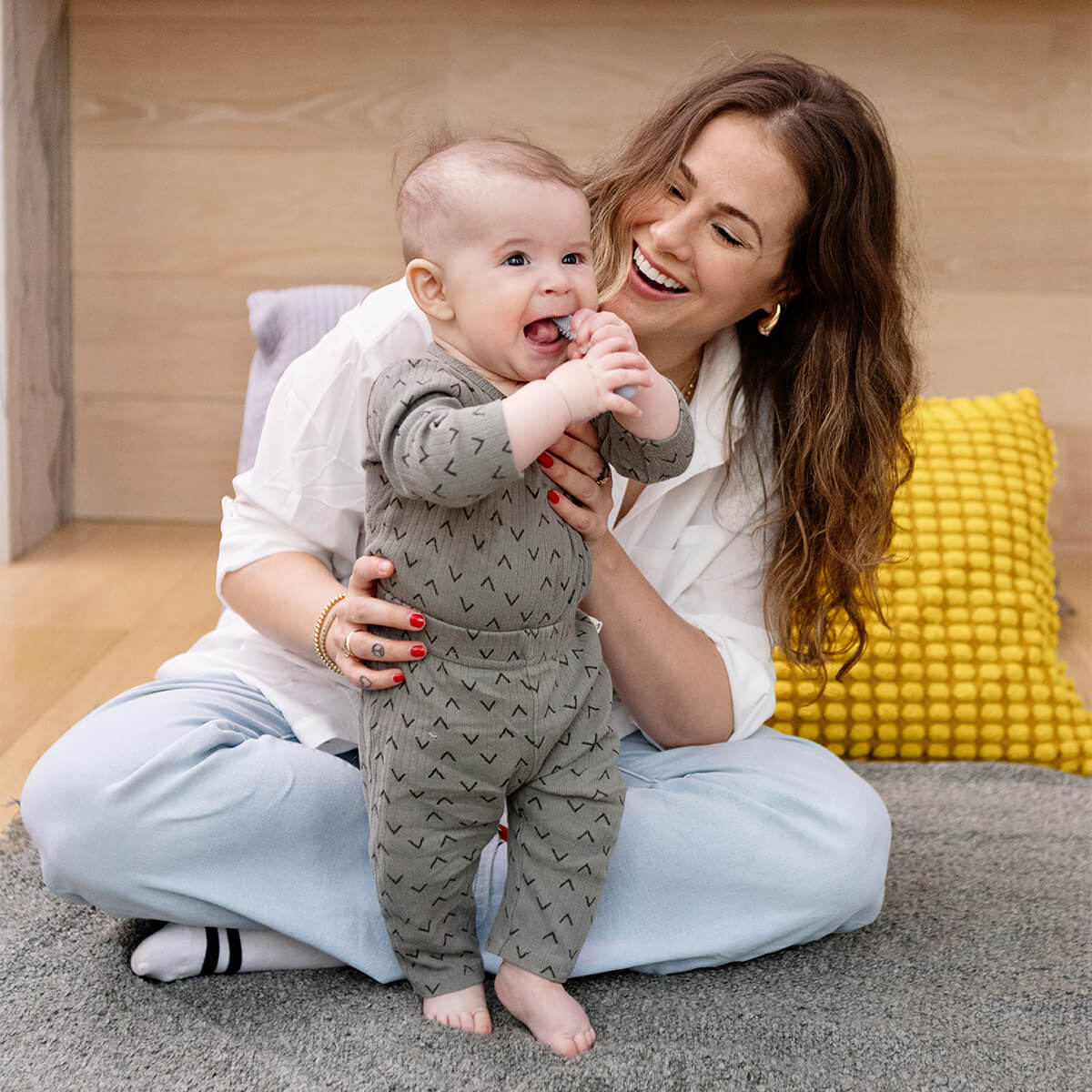 A woman helps a toddler stand while it puts the Pewter Baby-Led Toothbrush in its mouth.