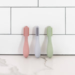 The Blush, Pewter, and Sage ezpz Baby-Led Toothbrushes leaning on a tile backsplash.