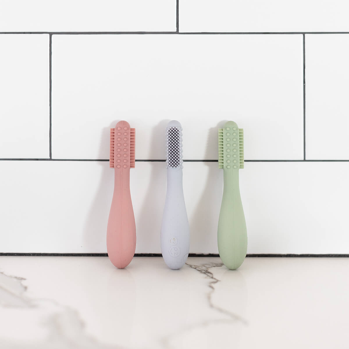The Blush, Pewter, and Sage ezpz Baby-Led Toothbrushes leaning on a tile backsplash.