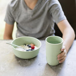 A child holds the Sage 14 oz Cup while sitting at a table next to a bowl of yoghurt.