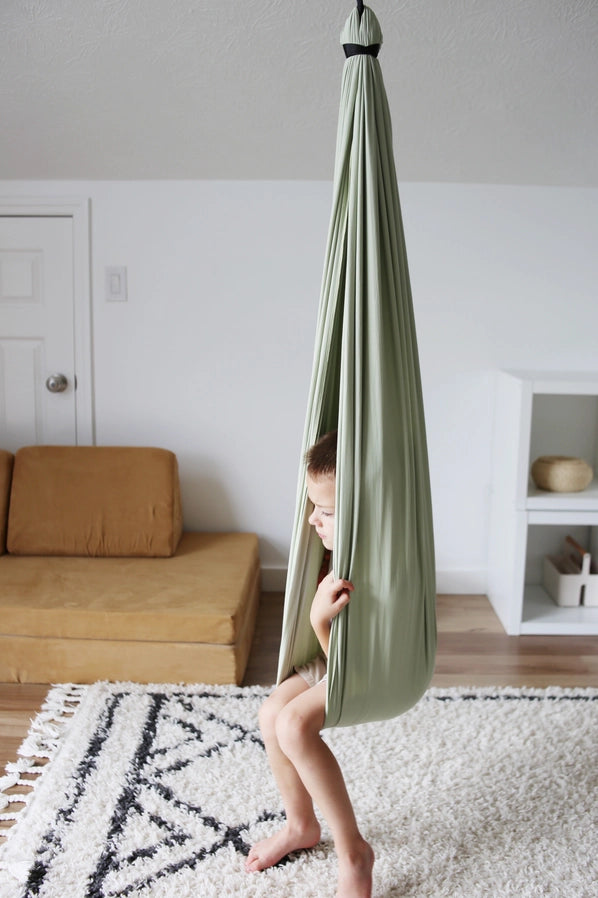 A child with light skin tone sits in the Desert Sage Sensory Swing with their legs dangling over the side.