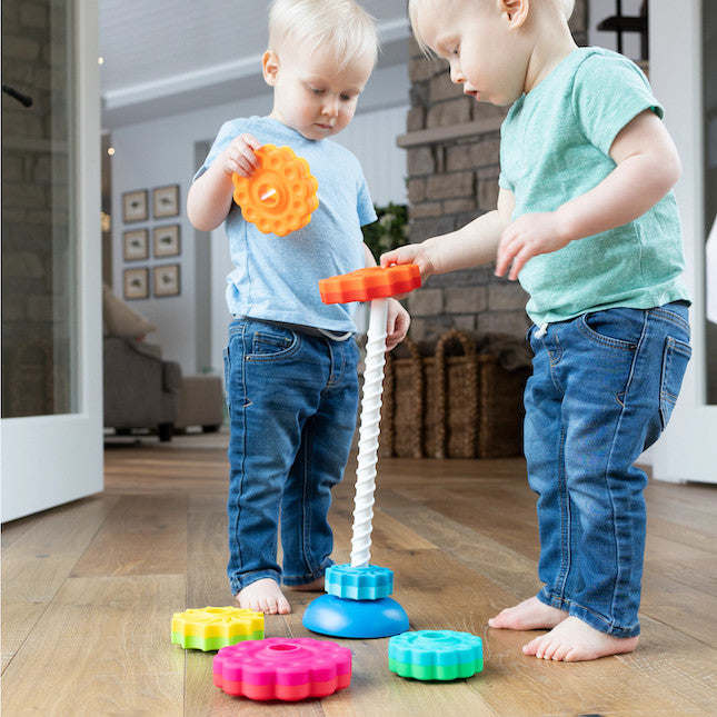 Two children with light skin tone and short blonde hair stand over the SpinAgain. One of them is putting a colorful piece down the spine and the other is waiting their turn.