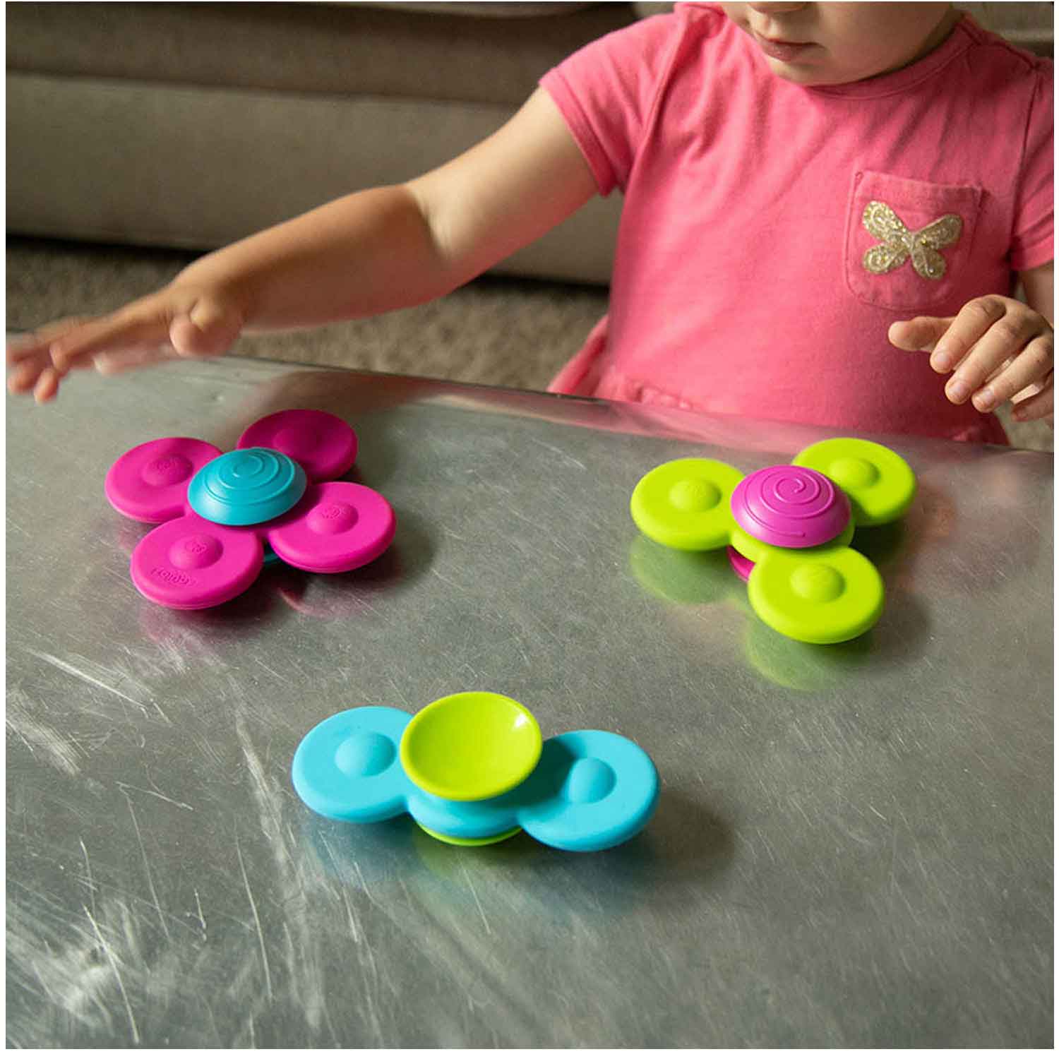 A baby with light skin tone sits at a metal counter and spins the Whirly Squigz.