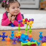 A toddler lays on the carpet and smiles at a precariously built figure composed of Squigz.