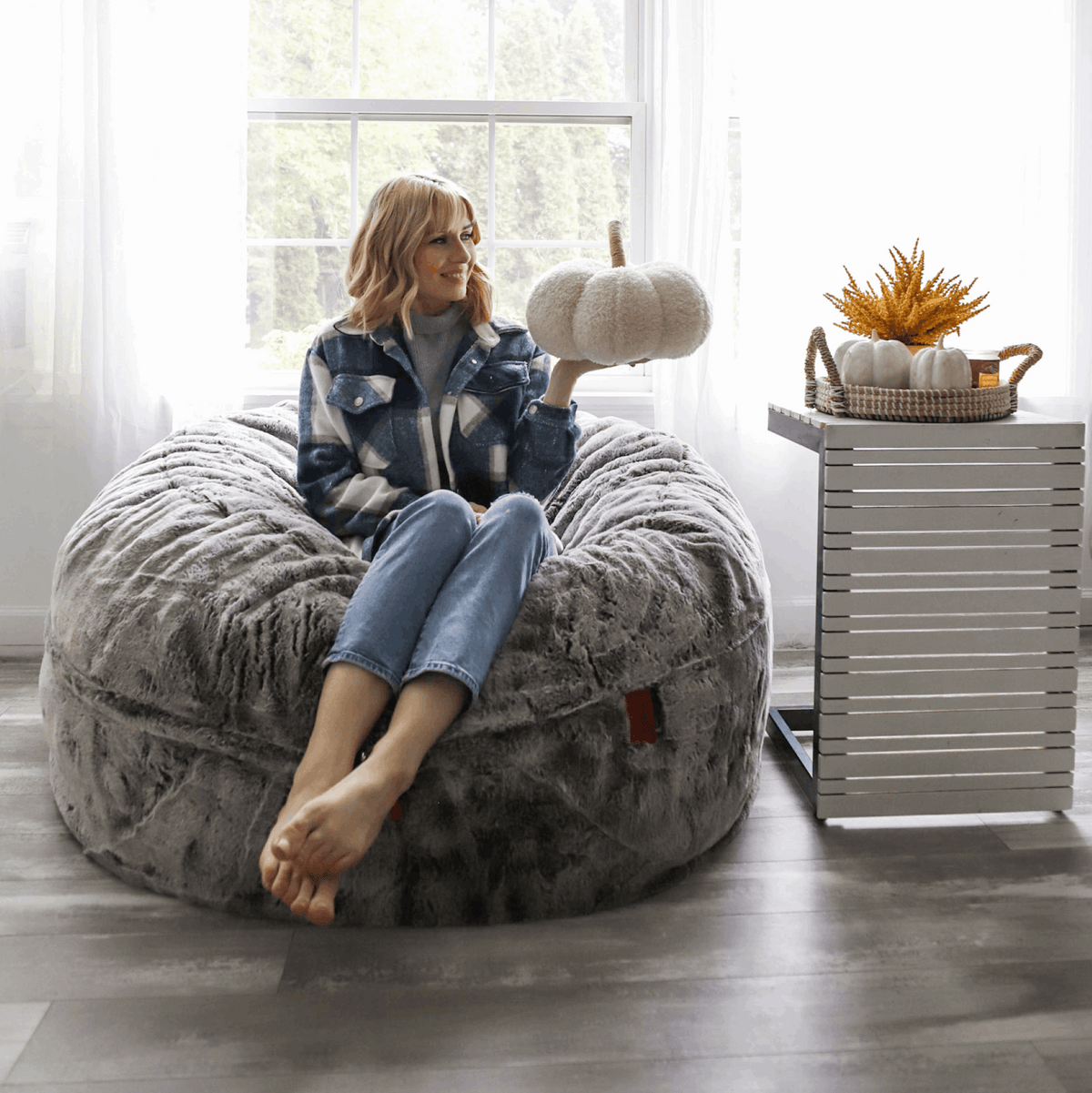 A woman sits on the Faux Chinchilla Bean Bag Convertible Bed as a chair and holds up a white decorative pumpkin.