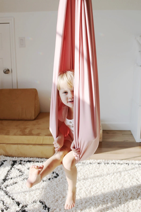 A child with light skin tone sits back in the Vintage Rose Sensory Swing with one leg stretched out in front of them and the other hanging down.