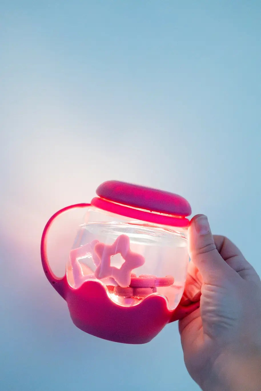 An adult hand holds up the pink Glo Pals Sensory Jar and Silicone Fun Filler while it glows.