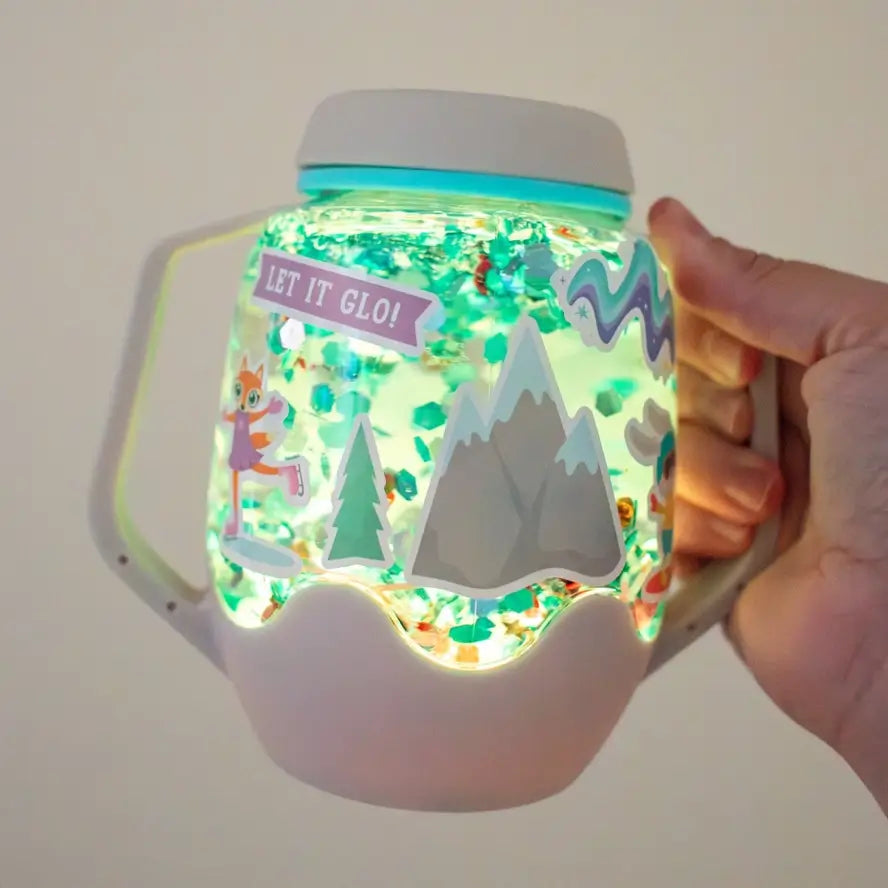 A hand holds up an illumnated white jar that is glowing from the inside. It is decorated with seasonal winter stickers and the inside has floating green glitter.