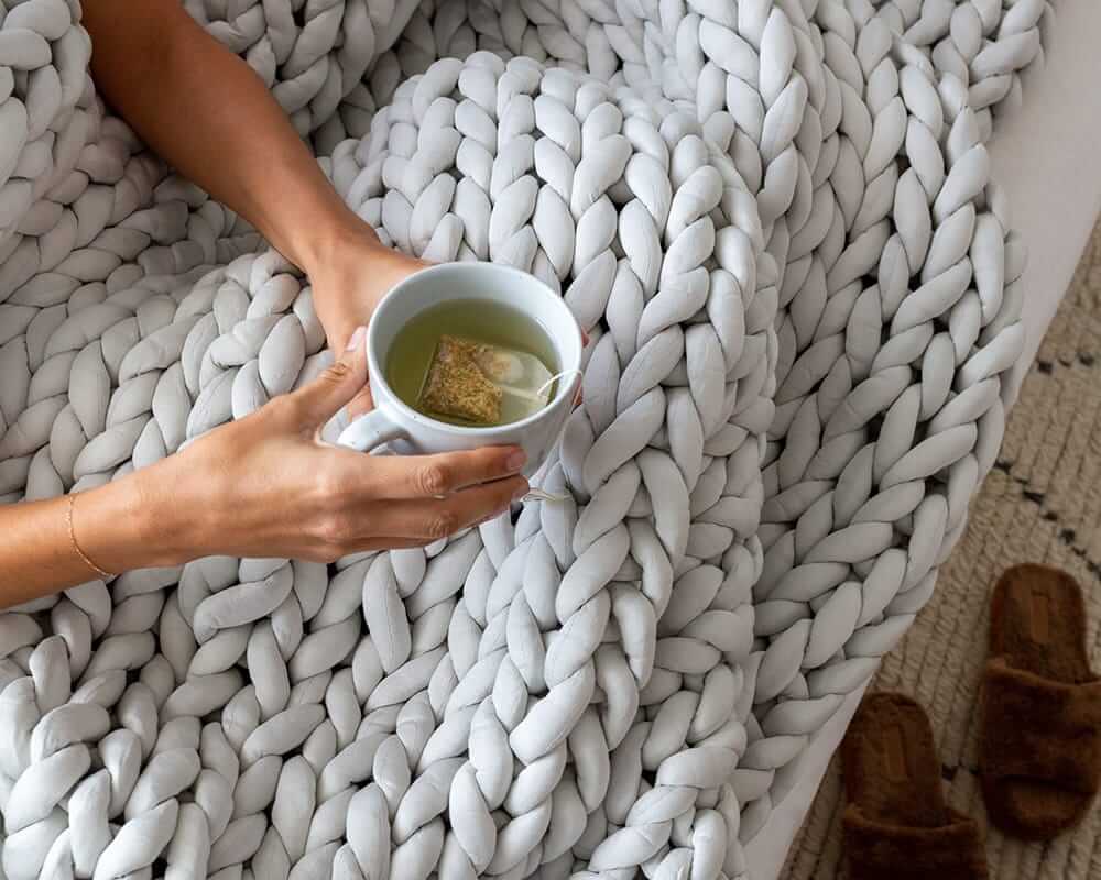 Two hands hold a full tea cup over the Soft Grey Chunky Knit Weighted Blanket spread over their lap.