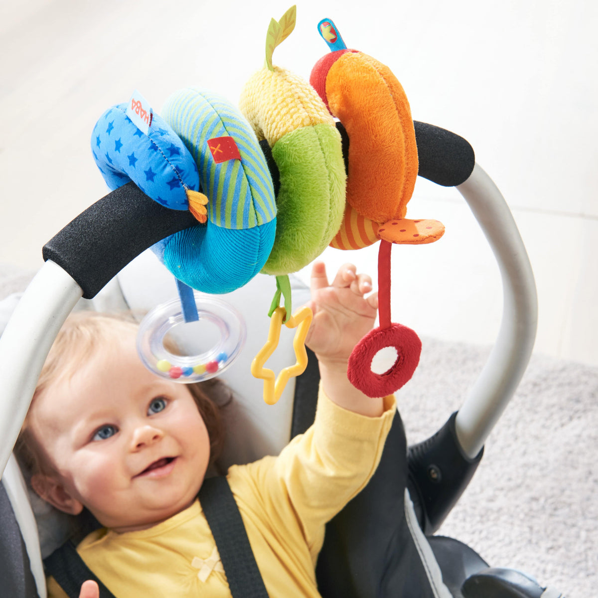 A baby in a car seat looks up at the handle which is decorated in hte Rainbow Activity Spiral Stroller & Car Seat Toy.