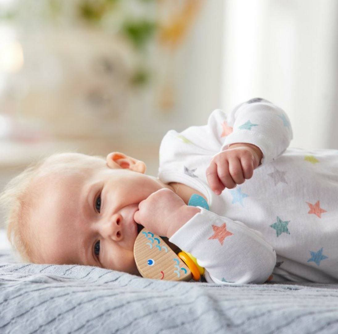 A baby lays on its side and puts the  Rattlefish Baby Wooden Rattle in its mouth.