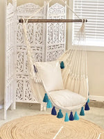 The Hammock Chair Stand with Turquoise Tassels hangs in a living room above a straw mat.
