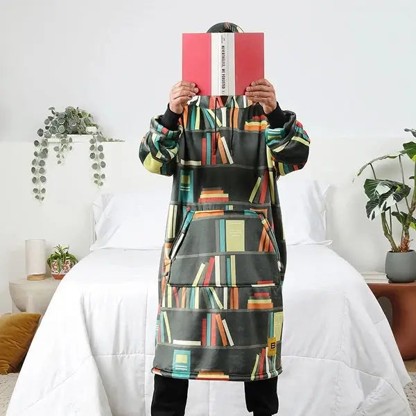 Someone stands in front of a bed wearing the Books Hideout Hoodie while holding an open book in front of their face.