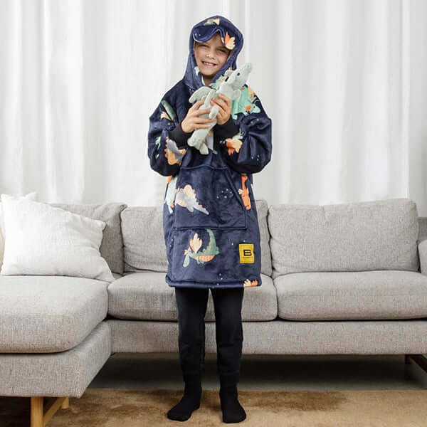 A child stands in front of a couch wearing the Dragons Kids Hideout Hoodie and holding a stuffed dragon.