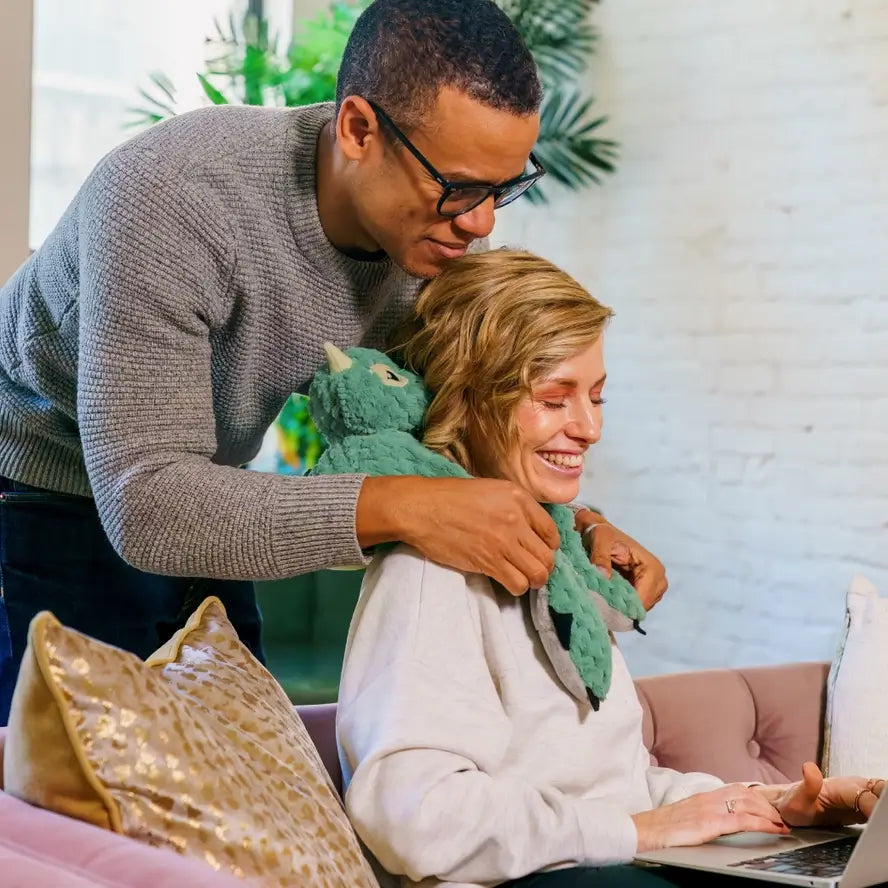 A man stands over the back of a woman on a laptop while adjusting Zephyr the Dragon over her shoulder while she smiles.
