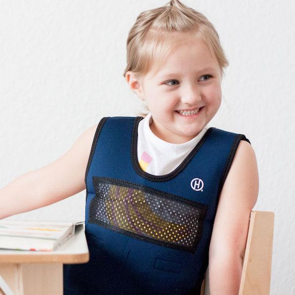 A child with light skin tone and blonde hair that is pulled back sits at a desk. They are wearing a Weighted Compression Vest over a short sleeved shirt.