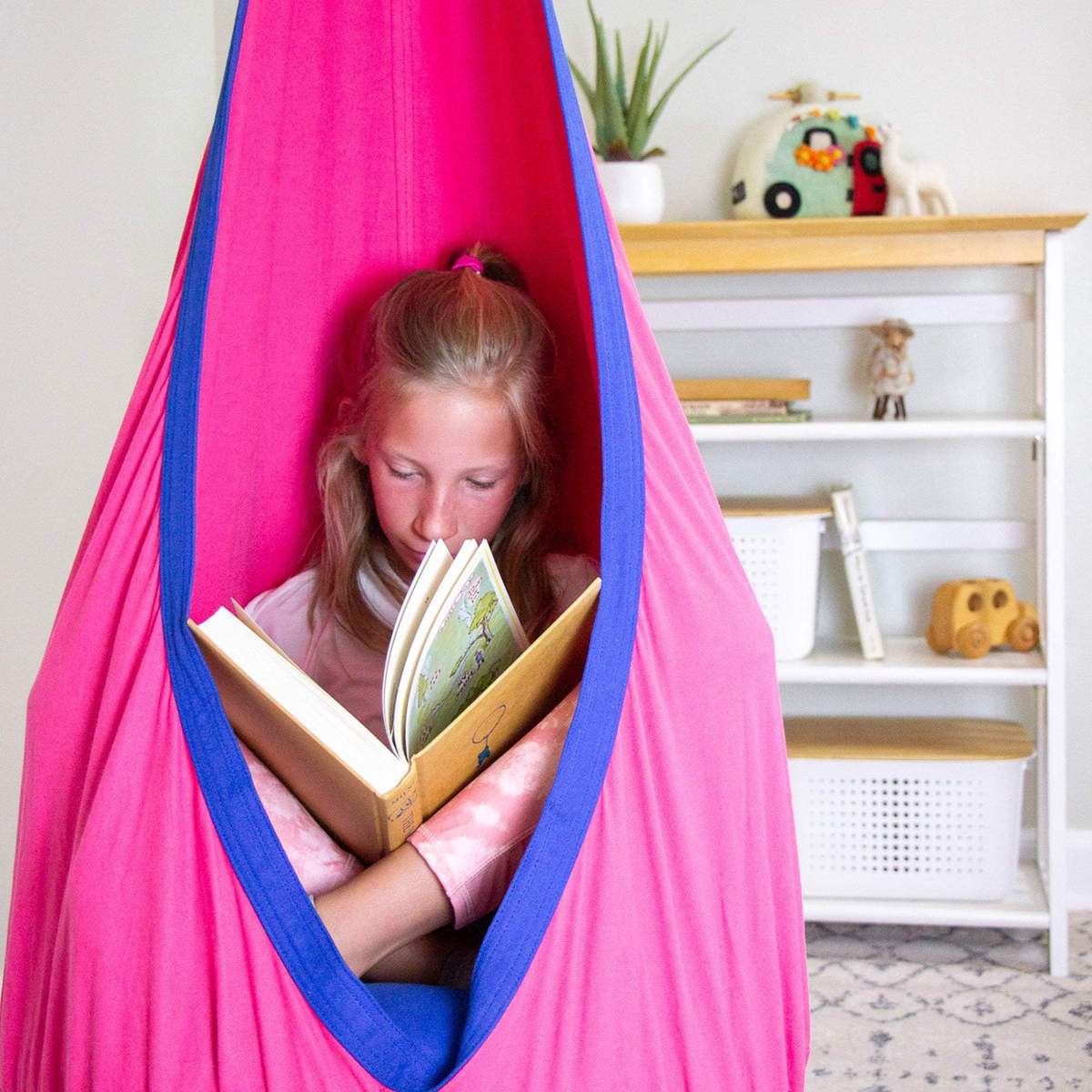 A child with light skin tone and blonde hair pulled back into a half ponytail sits in the pink Sensory Pod Swing. They are holding an open book and looking down at it.
