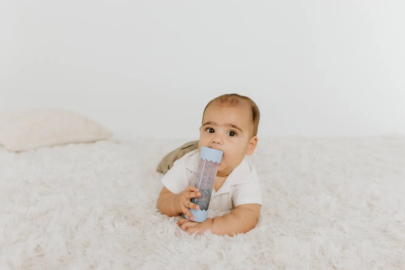 A baby on their tummy, holding the Soft Blue Baby Calm Down Bottle up to their mouth.