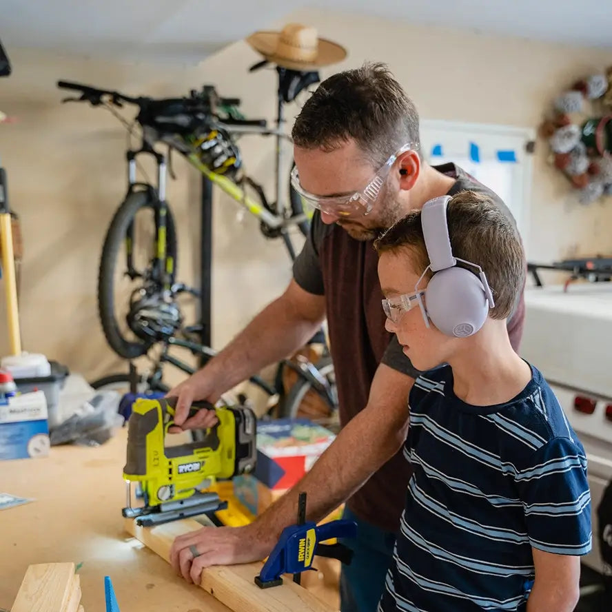 A pre-teen wears the Jlab Jbuddies Protect Kids Hearing Protect Earmuffs in Lilac while standing next to an adult using a power saw.