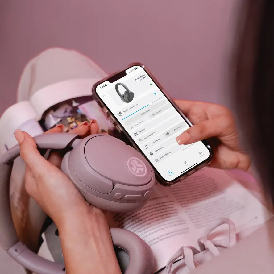 A model holds a pair of Mauve JLAB Jbuds Lux Active Noise Cancelling Headphones next to their phone while they pair them together.