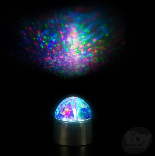 The Kaleidoscope Lamp with Batteries sits in the center of a dark area, projecting a multi-colored light show just above it.