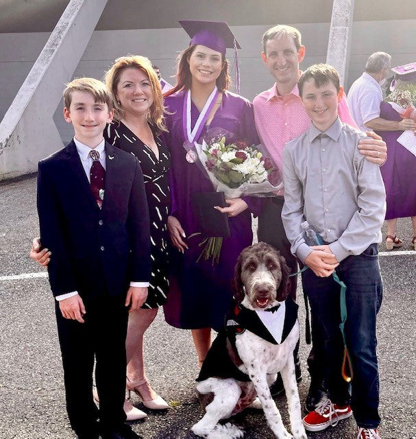 High school graduate holding flowers surrounded by mom, dad, and two brothers along with a large dog in a tuxedo